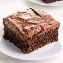A square-cropped photo capturing a tempting slice of whole wheat healthy chocolate cake placed on a plate, showcased on a clean white counter. The visual emphasizes the delightful presentation of the plant-based dessert against a minimalist backdrop.