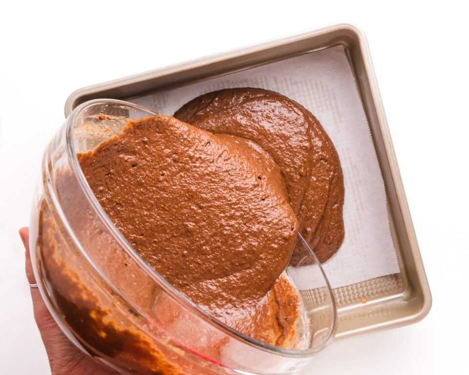 A step-by-step process shot capturing the pouring of chocolate cake batter into a square cake pan lined with parchment paper. The image illustrates the initial stage of creating the delectable whole wheat healthy chocolate cake, with the glossy batter smoothly filling the pan.