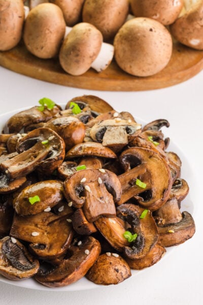 A plates of healthy sautéed mushrooms sits in front of a cutting board with raw mushrooms.
