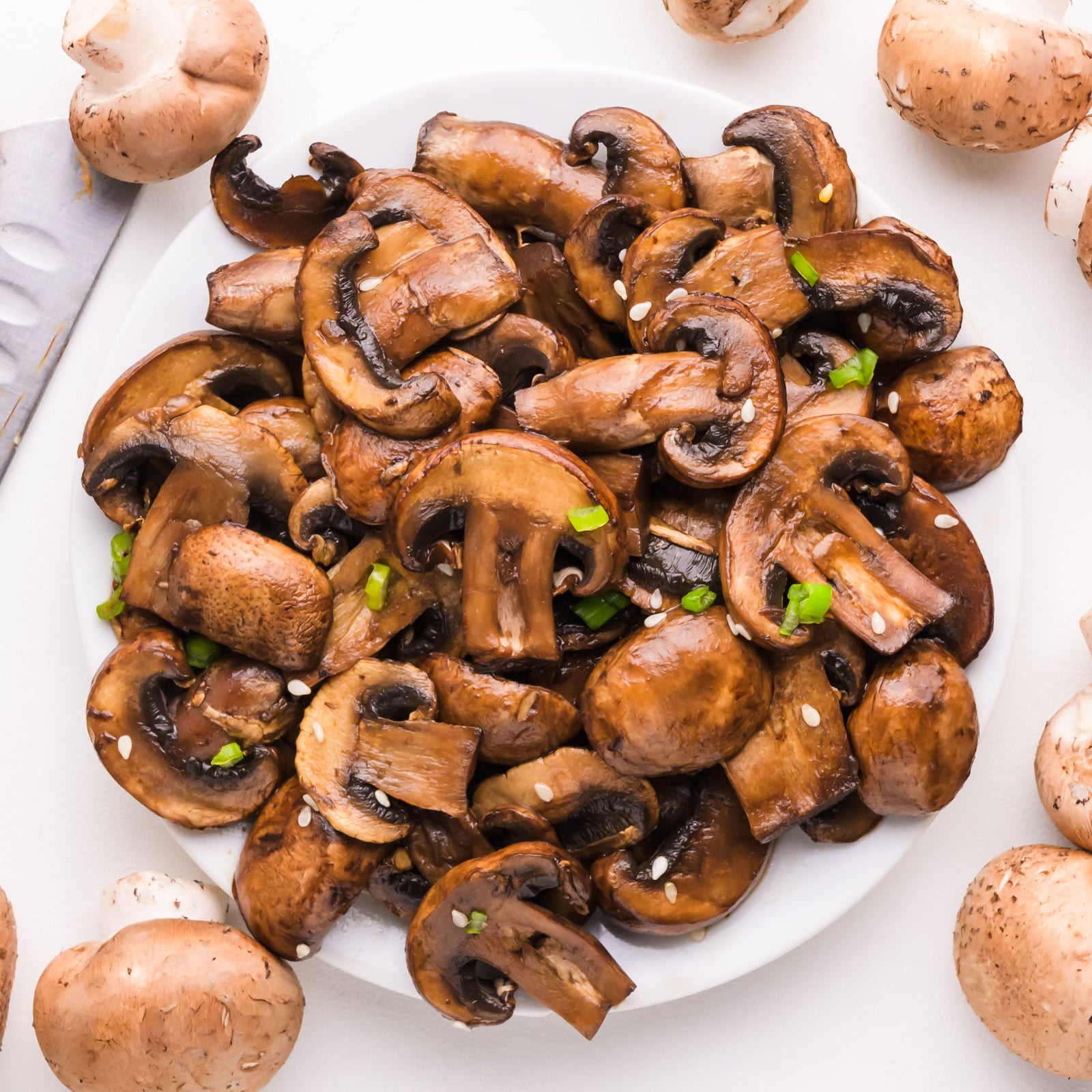 Looking down on sautéed mushrooms on a plate with whole mushrooms and a chef's knife around the plate.
