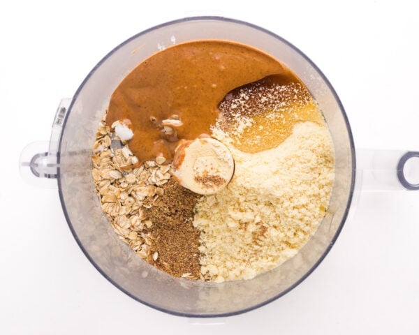 Looking down on ingredients in a food processor bowl, such as almond butter, oats, and more.