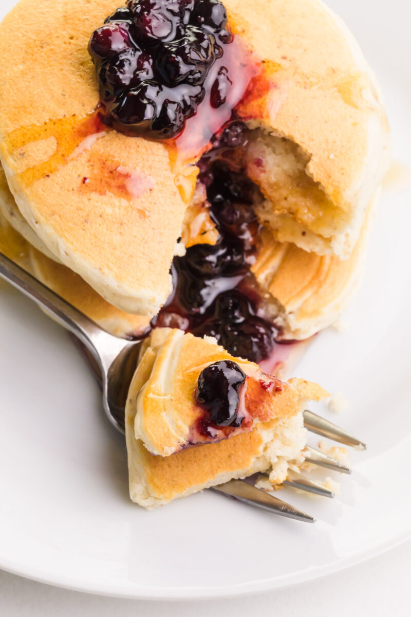 Looking down on pancakes with a bite taken out on a fork. There are blueberries and maple syrup on top.