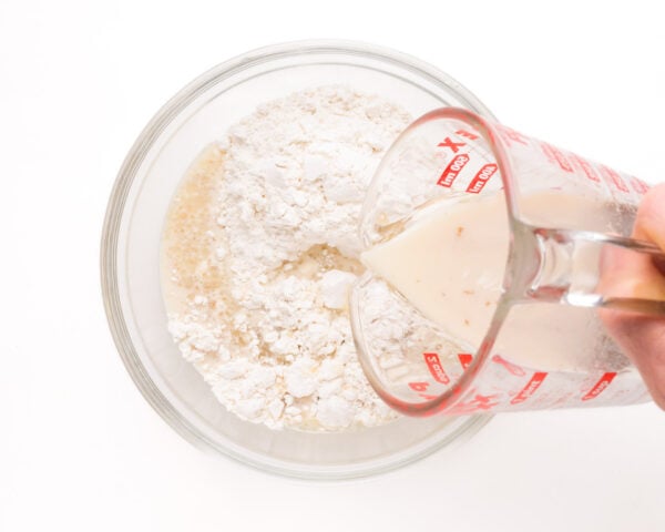 Milk mixture is being poured from a pyrex measuring dish into a bowl with flour.