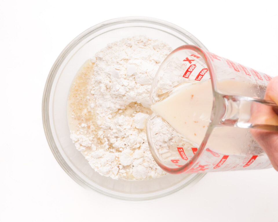 Milk mixture is being poured from a pyrex measuring dish into a bowl with flour.