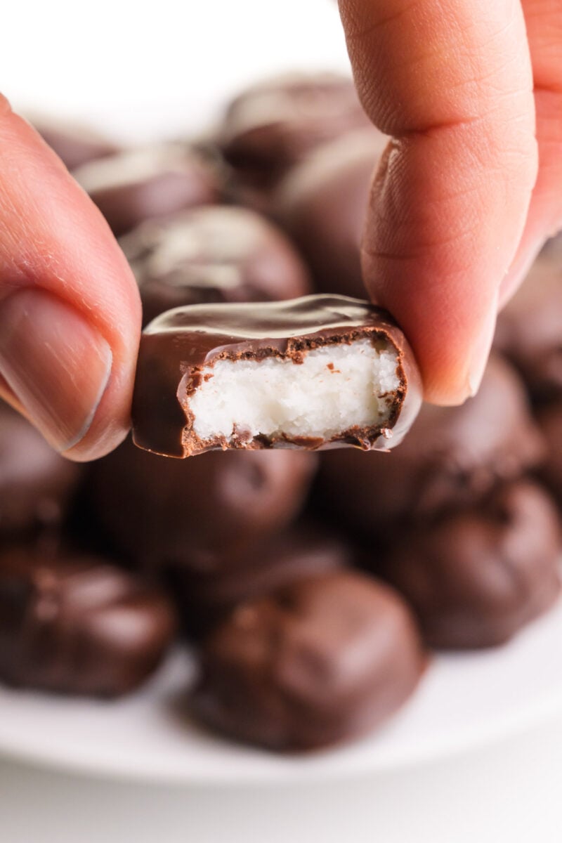 A hand holds a vegan junior mint with a bite taken out. There is a plate with more in the background. 