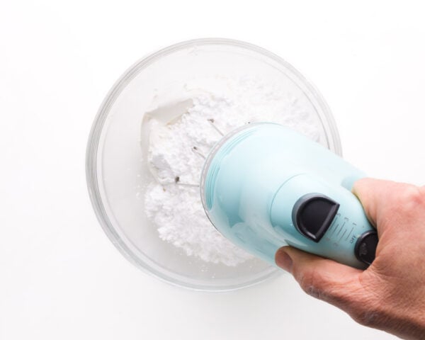 A hand holds a blue mixer, beating creamy white mixture in a mixing bowl.