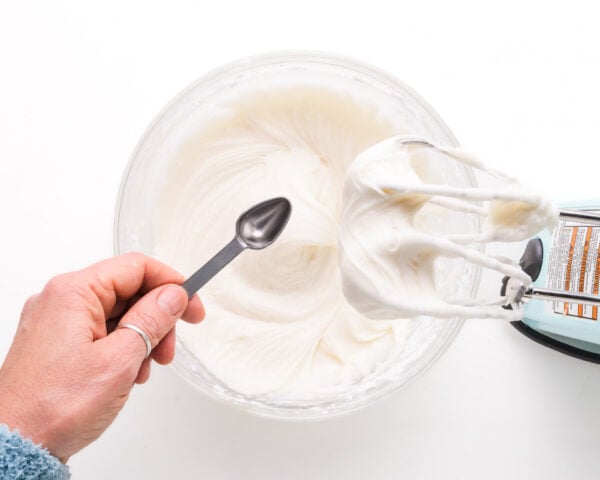 A hand pours in mint extract into a bowl with creamy white mixture sitting next to an electric mixer.
