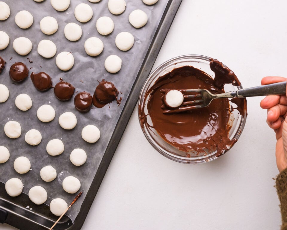 A hand dips mint patties into melted dark chocolate. There is a pan with both dipped and plain patties.