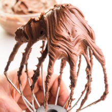 Chocolate ganache frosting is filling up a stand mixer's whipping attachment.
