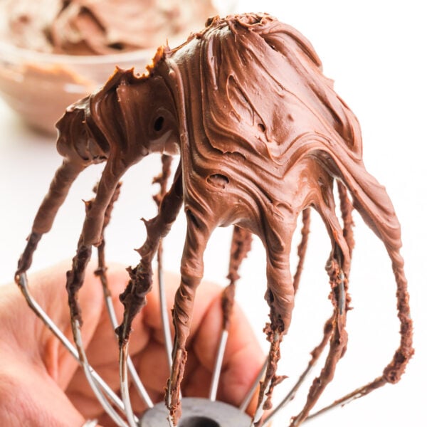 Chocolate ganache frosting is filling up a stand mixer's whipping attachment.