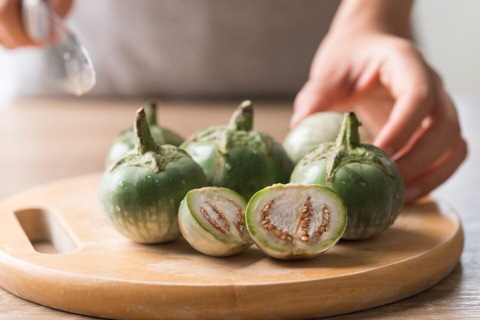 Hand holding knife preparing to cut green eggplants on a wooden cutting board.