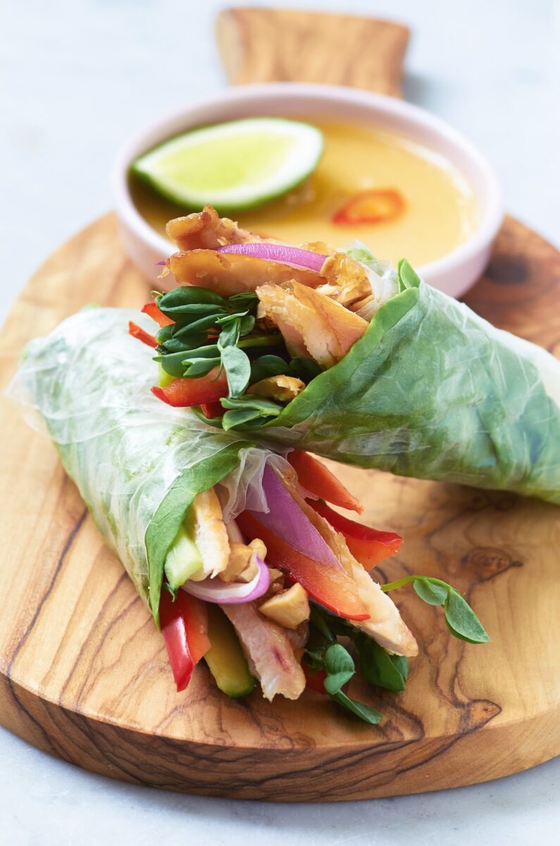 Two spring rolls are on a cutting board with a bowl of sauce in the background.