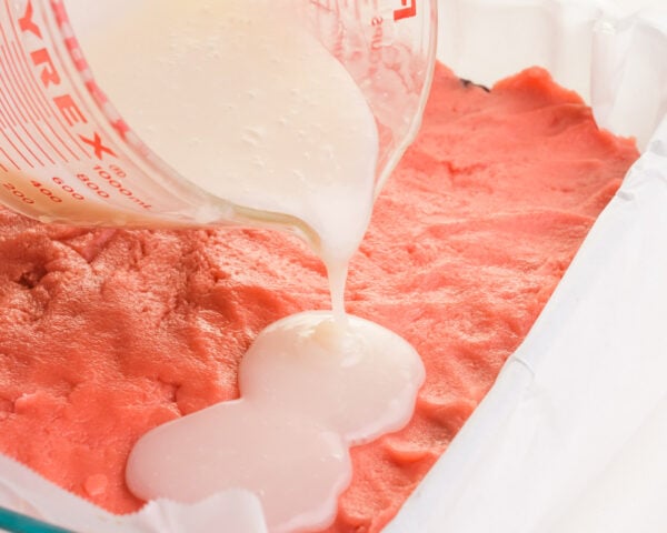 A coconut milk mixture is being poured into a pan with pink strawberry cake batter.