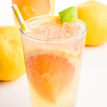 A closeup of a glass of grapefruit green tea with grapefruit wedges and a paper straw. There are whole fruits in the background and another glass.