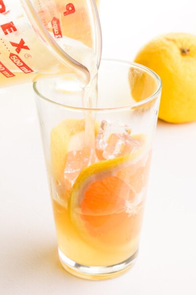 A green tea mixture is being poured into a glass with ice and grapefruit slices. There is a grapefruit in the background.