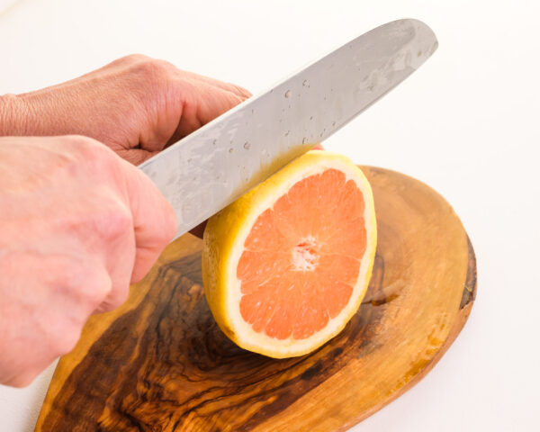 A hand holds a knife, cutting a grapefruit on a cutting board.