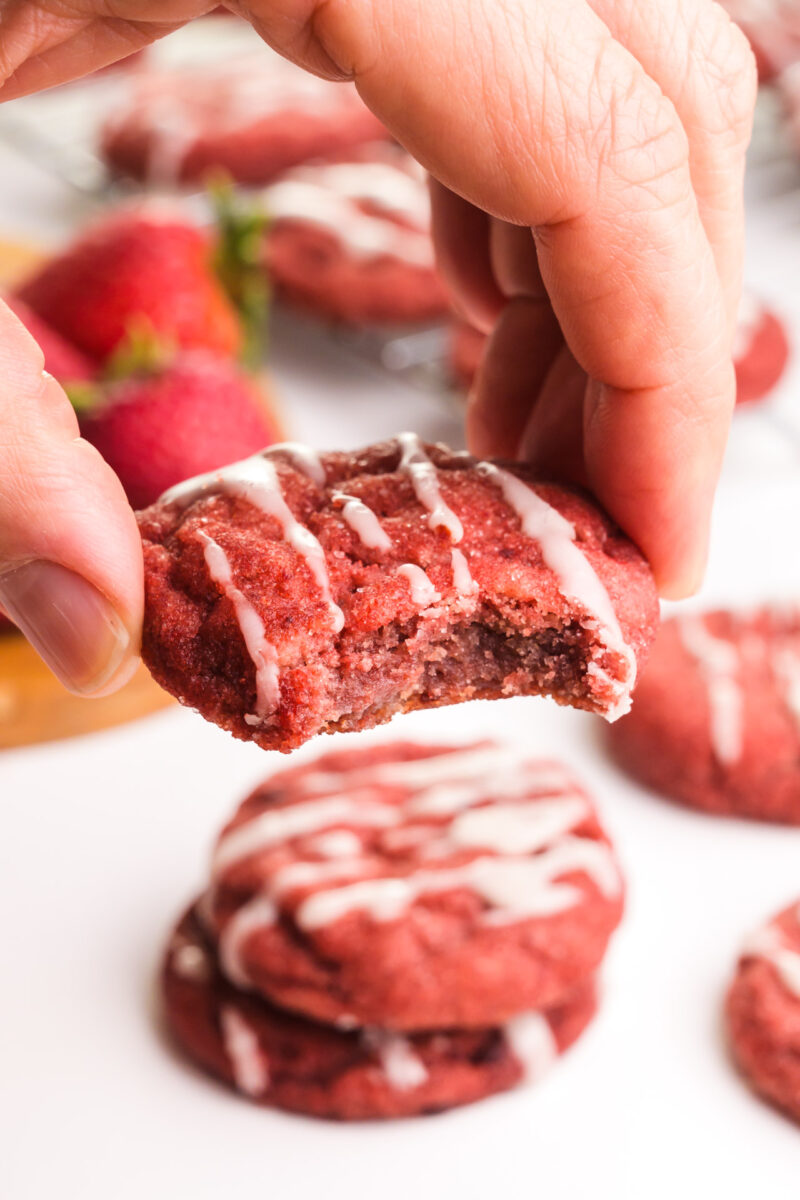 A hand holds a vegan strawberry cookie with a bite taken out. It hovers over more cookies in the background.