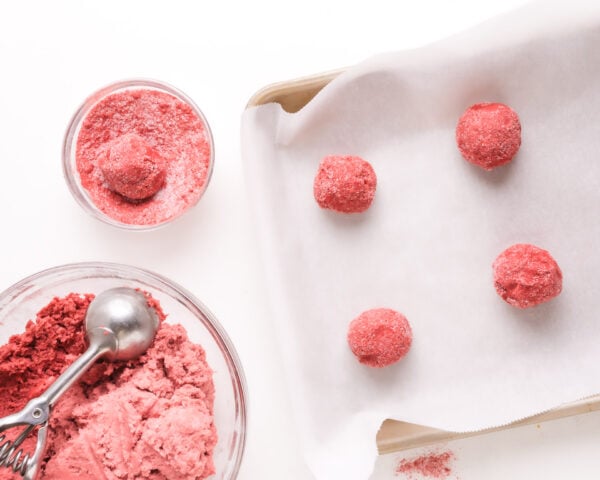 Looking down on cookie dough balls on a pan and more being rolled in a strawberry sugar mixture.