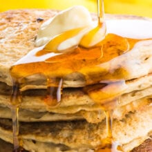 Maple syrup is being drizzled over banana pancakes with a pat of melty butter on top.