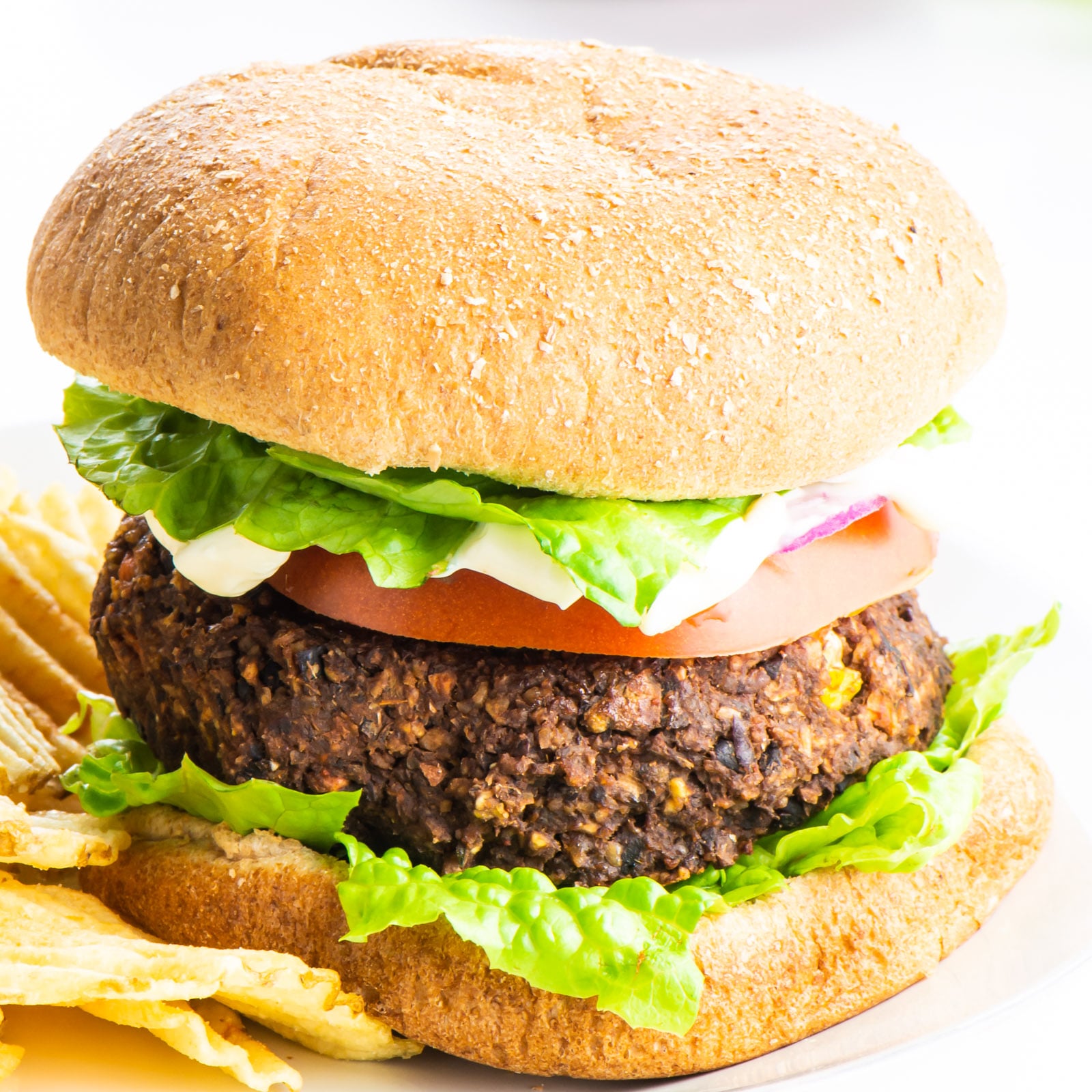 A bean burger is on a bun and topped with veggies like tomatoes and lettuce. It also has mayo on top. It sits beside potato chips.