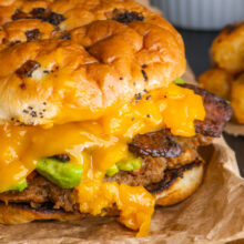 A closeup of a veggie burger on a bun shows lots of melted cheese, avocado slices, and veggie bacon on top.