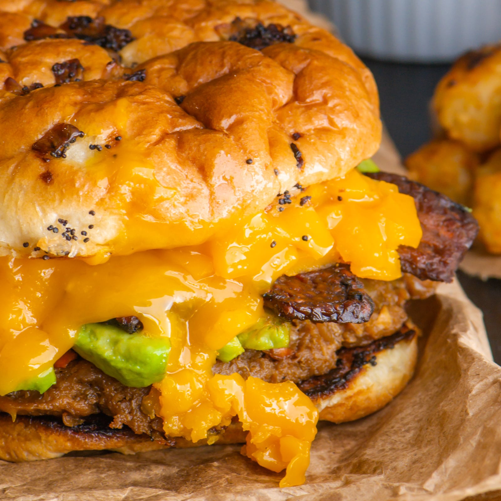 A closeup of a veggie burger on a bun shows lots of melted cheese, avocado slices, and veggie bacon on top.