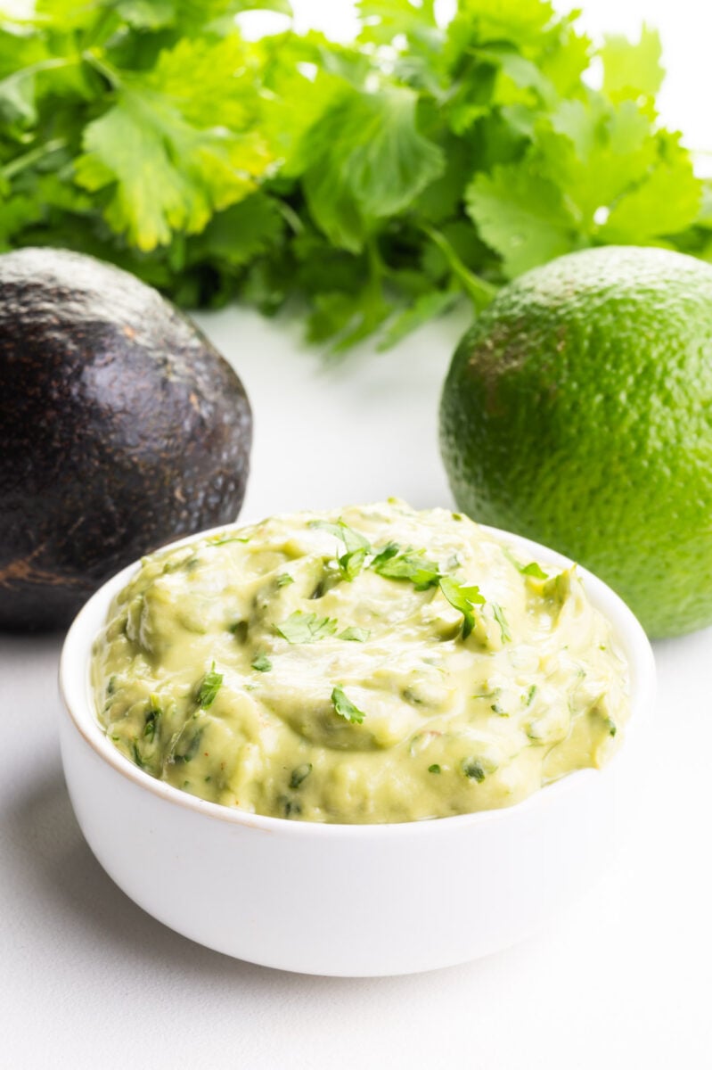 A bowl of avocado sauce sits in front of a whole avocado, a lime, and herbs.