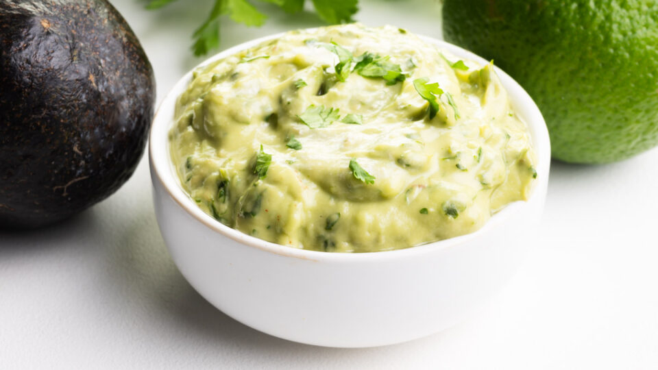 A closeup of a small bowl of avocado cilantro lime rice. There is an avocado, fresh herbs, and a lime in the background.
