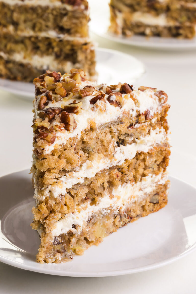 Layered cake with cream and nuts, served on a white plate, with additional cake slices in the background.
