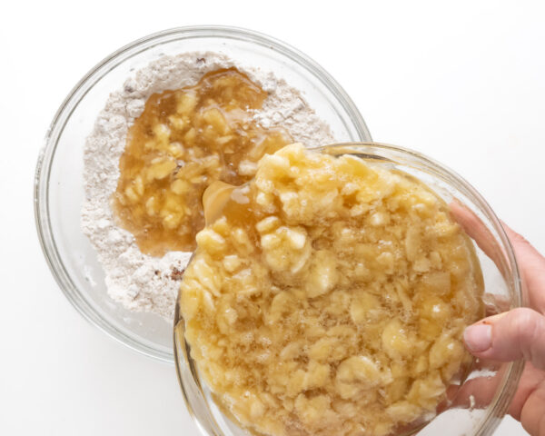A bowl of liquid cake batter mixture is being poured into a bowl with dry ingredients, such as flour.