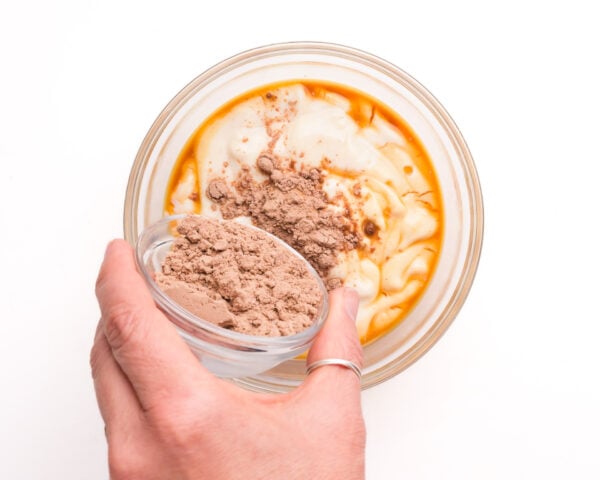 A hand pours chocolate protein powder into a bowl with other ingredients, like yogurt and vanilla extract.