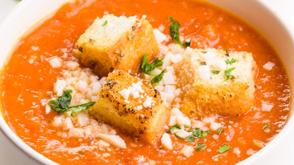 A bowl of tomato soup has croutons, herbs, and parmesan cheese on top.