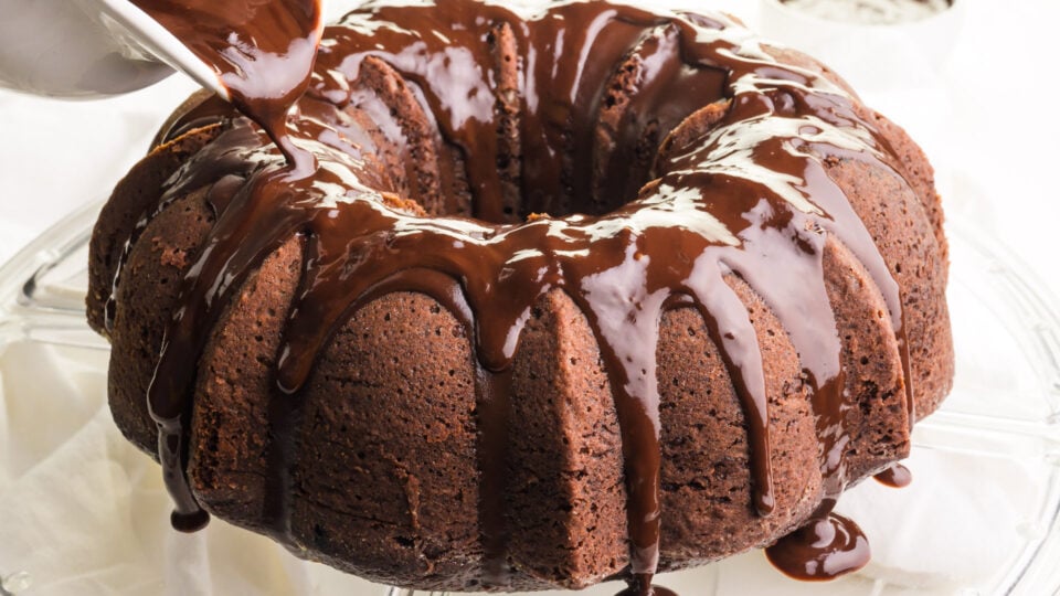 Chocolate sauce is being poured from a pitcher onto a chocolate Bundt Cake.