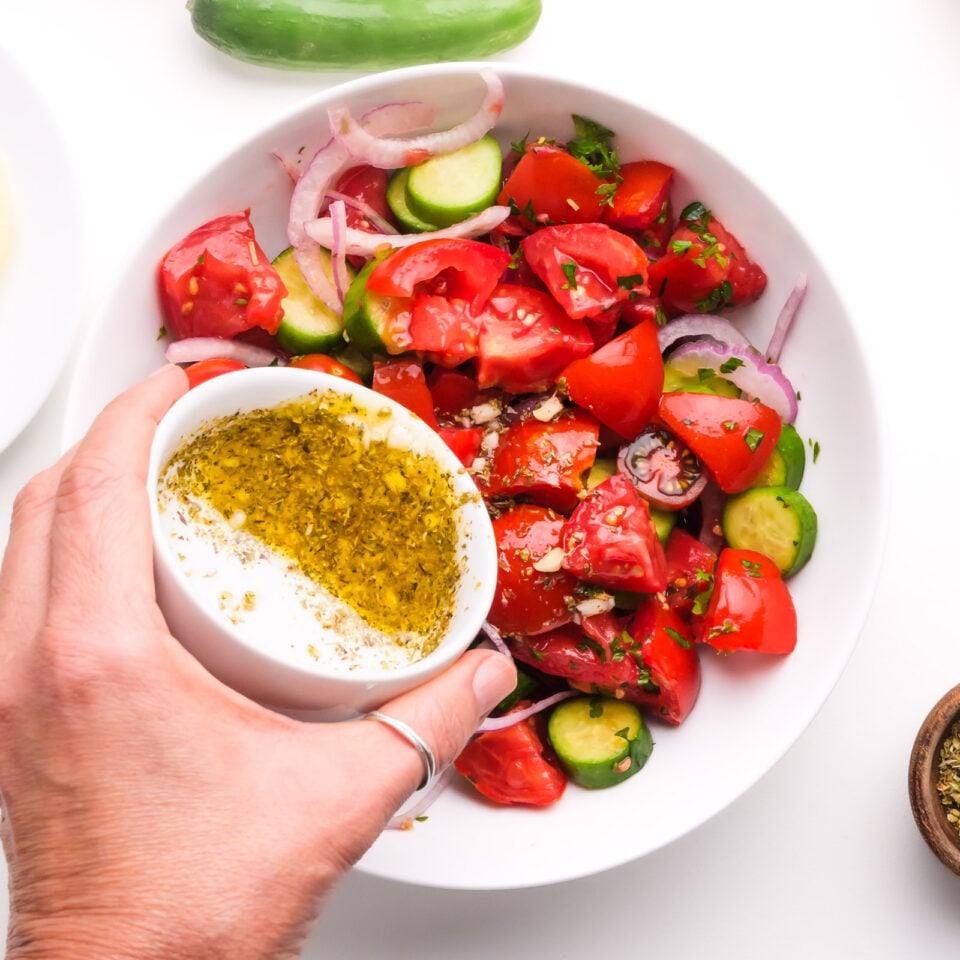 Dressing is being poured over tomato cucumber salad in a bowl.