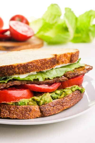 Layered sandwich with lettuce, tomatoes, avocado, and crispy bacon on toasted bread, with fresh vegetables in the background.