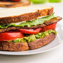 Close-up of a sandwich with layers of lettuce, tomatoes, guacamole, and a crispy exterior served on a plate.