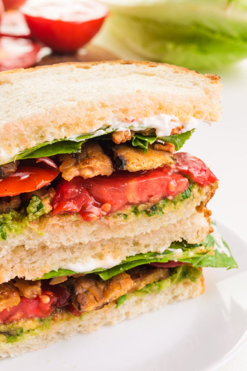 A vegan BLT sandwich is cut in half and stacked on top of each other, revealing layers of veggie bacon, sliced tomatoes, and more.