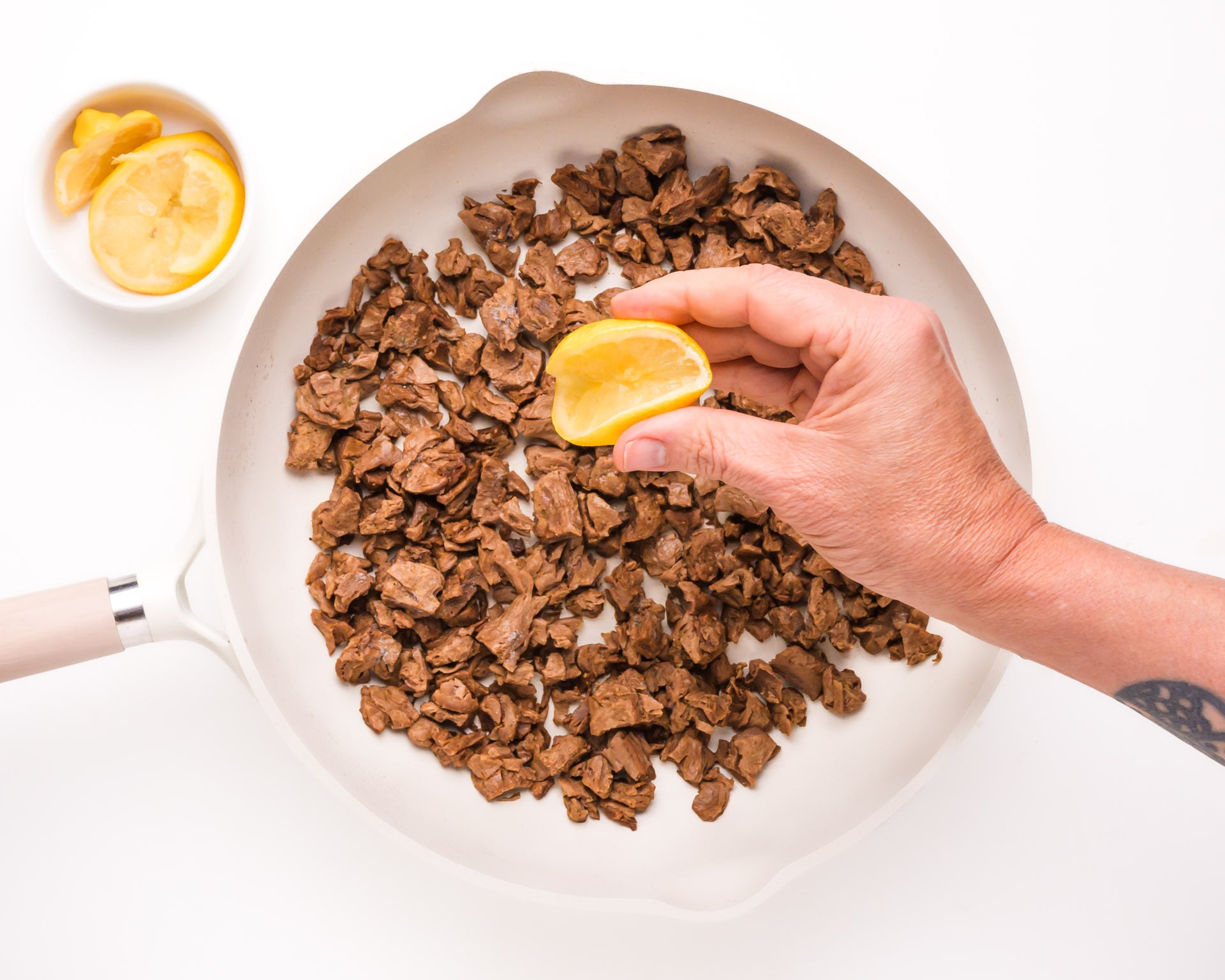 A hand squeezes a lemon over a pan with vegan steak pieces. A bowl with more lemon slices sits beside the pan.