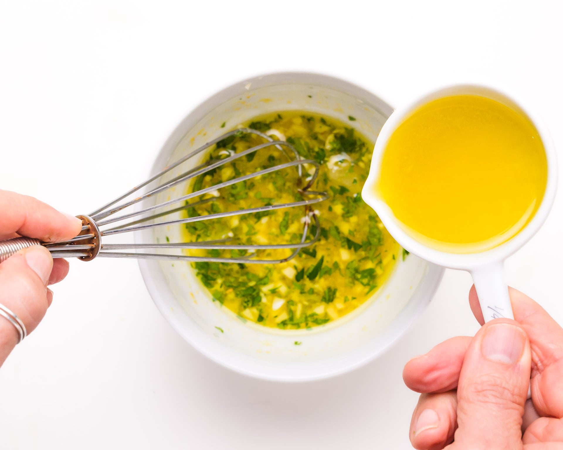 Olive oil is being drizzled over a bowl with an herb/lemon juice mixture as it is being whisked.