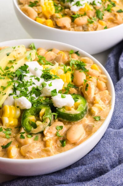 a bowl of white bean chili topped with sliced jalapenos, corn, sour cream and parsley