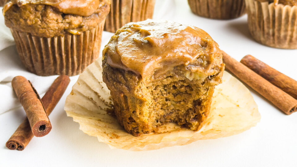 An iced muffin with a bite taken out sits in front of more muffins, a cup of coffee, and cinnamon sticks.