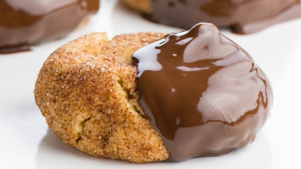 A snickerdoodle cookie dipped half-way in shiny chocolate sits on a white counter.