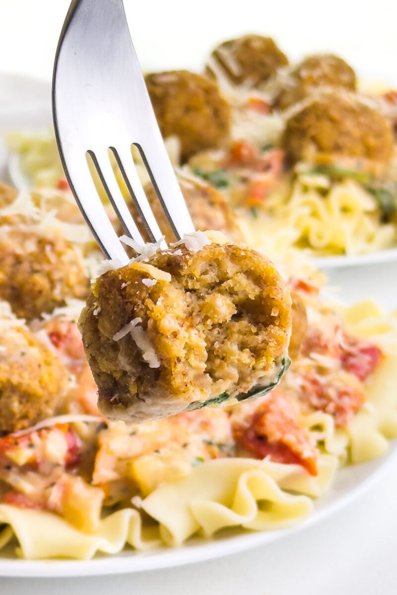 A fork holds a vegan chicken meatball with plates of the meatballs in the background.