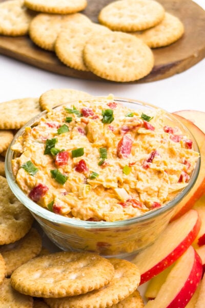 A dish of cheese spread sits around crackers and apple slices. There is a wooden cutting board with more crackers in the background.