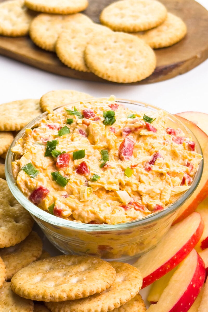 A dish of cheese spread sits around crackers and apple slices. There is a wooden cutting board with more crackers in the background.