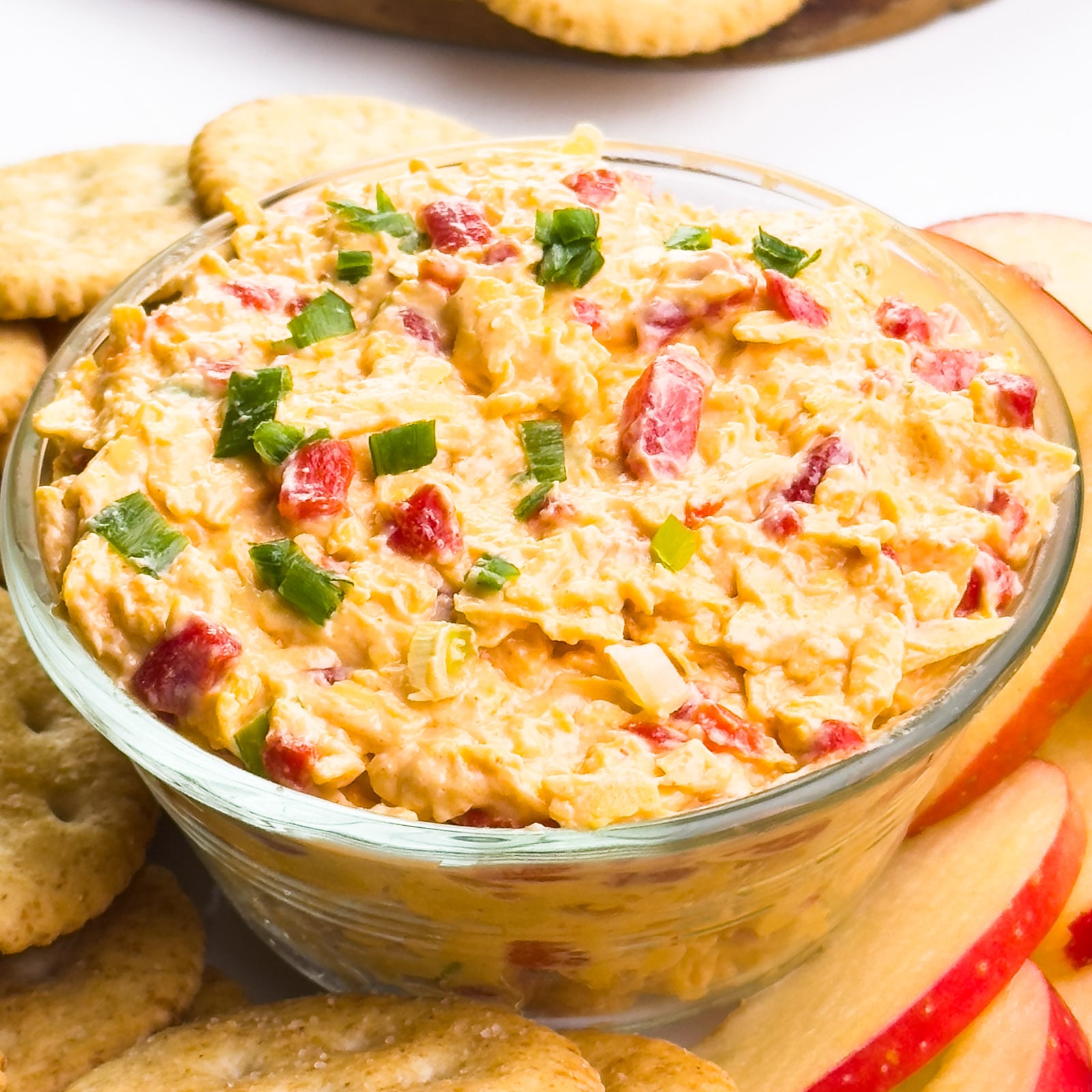 A bowl of pimento cheese sits on a plate with crackers and apple slices.