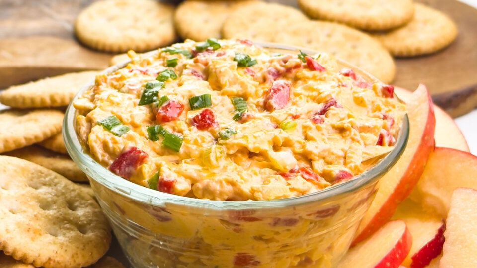 A bowl of pimento cheese spread sits amongst crackers and apple slices.