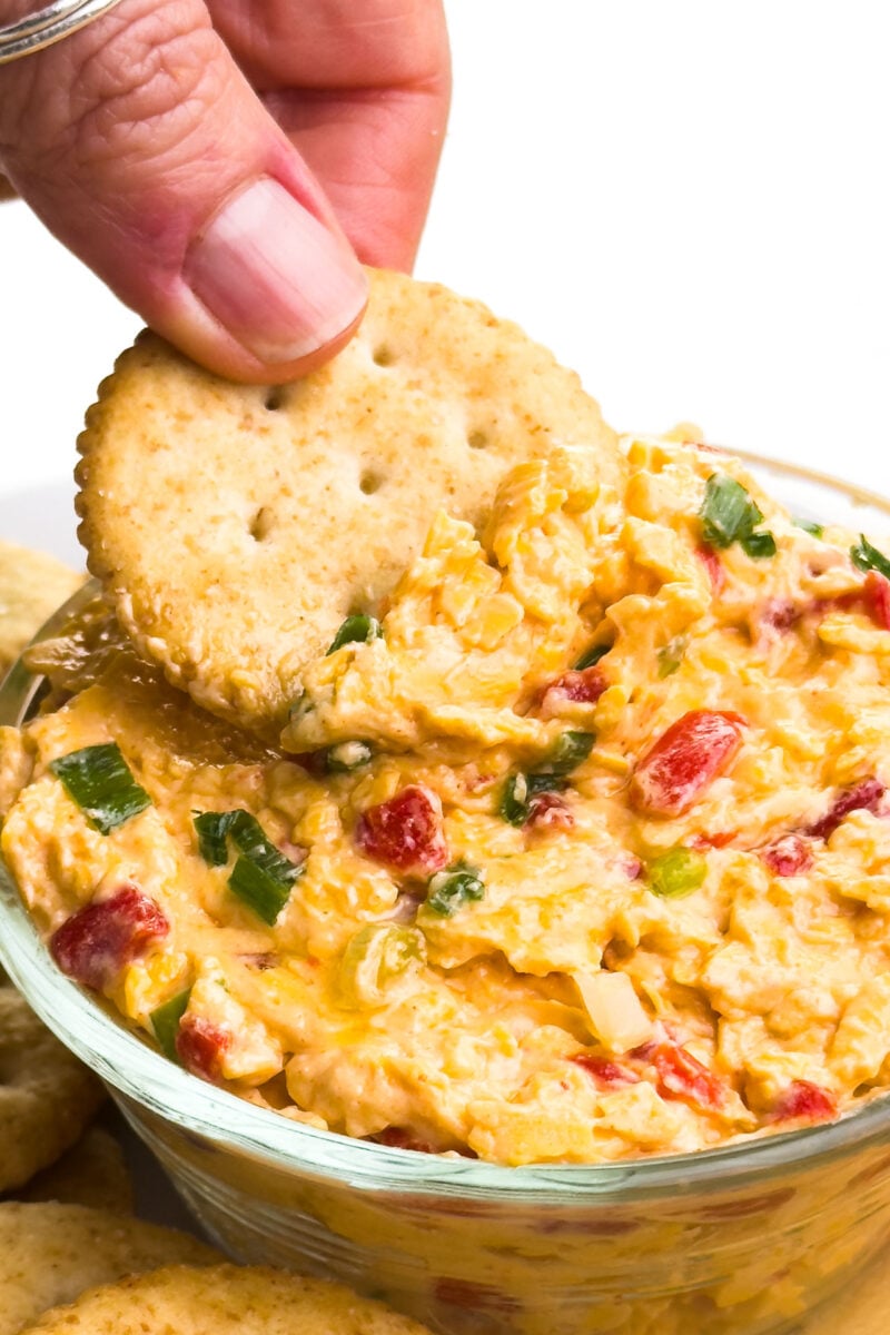 A hand dps a cracker into a bowl of vegan pimento cheese spread.