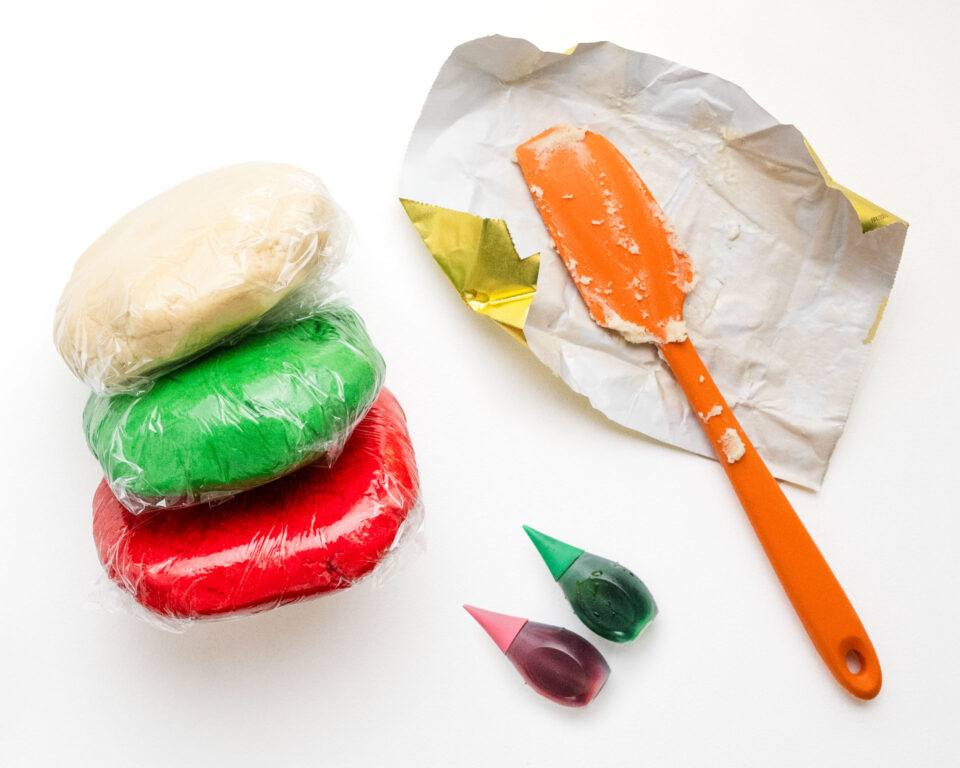 Three discs of colored cookie dough are in plastic wrap next to food coloring and a spatula.