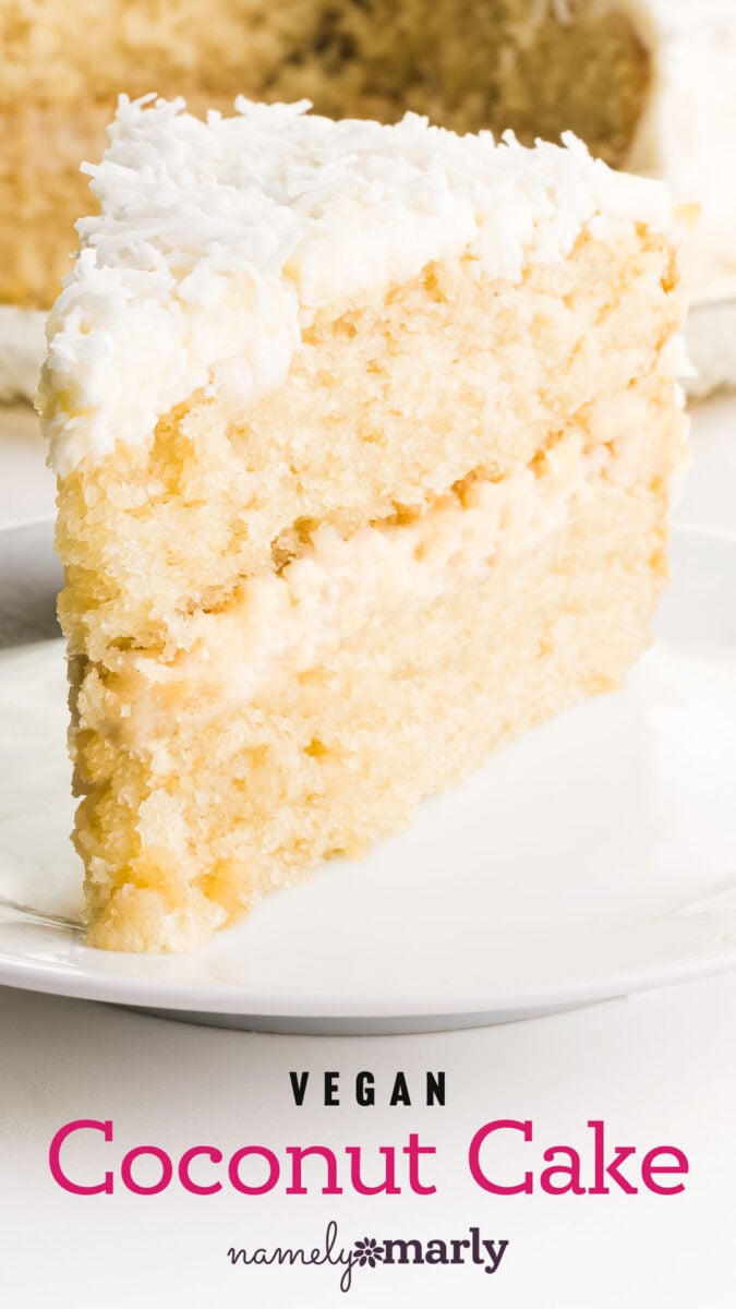 A slice of vegan coconut cake with white frosting and shredded coconut on top, served on a white plate.